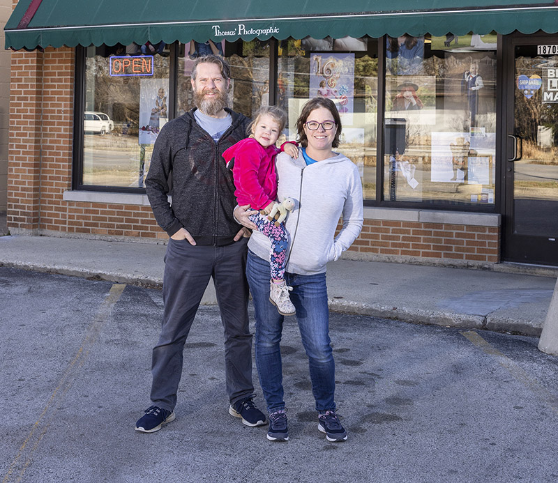 Thomas Photographic owners Colin, left, and Angela Thomas with their daughter, Elloren, in front of their current location at 18705 Dixie Highway. The couple currently is working to remodel space in a storefront across the street where they plan to move to in coming weeks. (Provided photo)