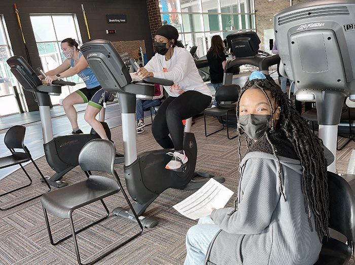 While Kristen Salkas, left, and Cori Williams ride stationary bicycles, Fitness Club volunteer Kamilah Muhammad sits nearby keeping track with her timesheet. (Nick Ulanowski/H-F Chronicle)