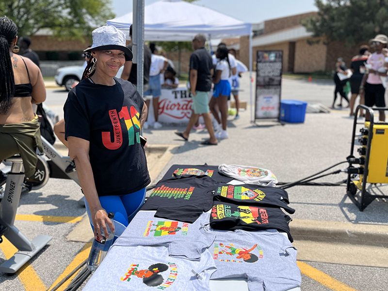 Cycling instructor Alicia Moore standing at booth behind exercise cycles at the HF Juneteenth Festival. (Nuha Abdessalam/H-F Chronicle)