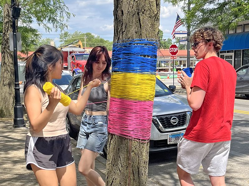 From left, Anly Flores, Anna Arruvo and Sullivam Matthys wrap blue, yellow and pink yarn, the colors of the pansexual pride flag, around a tree in a yarn bombing project. (Nick Ulanowski/H-F Chronicle)