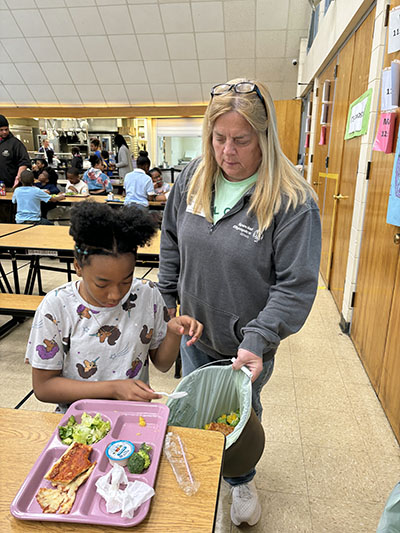 Churchill School student Senaya Marshall puts her peaches into the composting bucket for parent volunteer Patty Welsh. (Marilyn Thomas/H-F Chronicle)