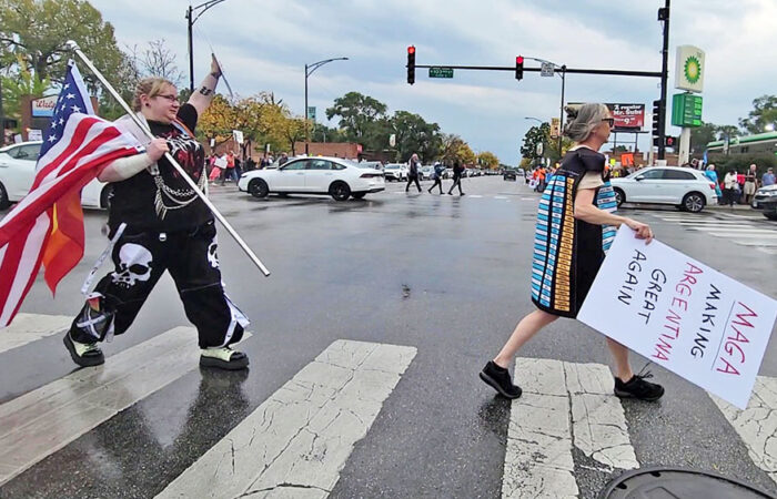 Two protesters cross Western Avenue during the No Kings demonstration on Oct. 18, one holding a sign that reads "MAGA making Argentina great again," a reference to the recent news that the Trump administration was sending billions of dollars in aid to the South American country, and one carrying an American flag. (Eric Crump/H-F Chronicle)