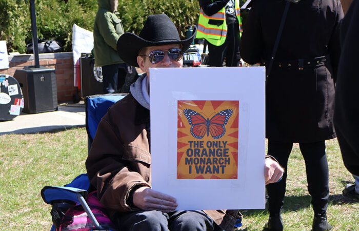 A protester holds a sign reading “The only orange monarch I want” while seated near the Homewood Veterans Memorial during Saturday’s rally. (Nuha Abdessalam/H-F Chronicle)
