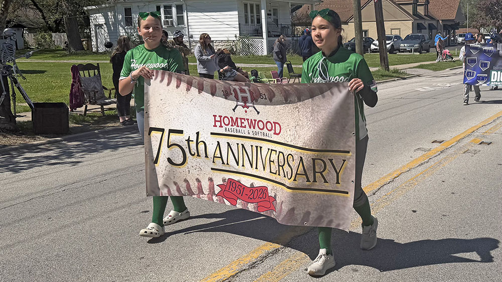 Two softball players carry a banner in the opening day parade recognizing Homewood Baseball and Softball's 75th anniversary. (Nick Ulanowski / H-F Chronicle)